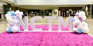 A picture of a conference center with a large set of letters spelling NCTM on a pink carpet, with balloons on each side.